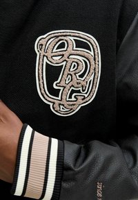 Sort varsity-jakke med beige broderede sammenflettede "D" og "R" bogstaver på brystet, ribbet kant med beige, hvid og sort striber.