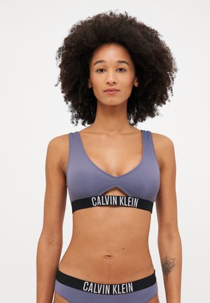 Soutien-gorge de sport violet cannelé avec une bande élastique noire portant le logo "CALVIN KLEIN". Décolleté en V profond et détail découpé à l'avant.