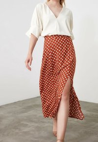 Gonna a pois marrone ruggine con pois bianchi, caratterizzata da uno spacco alto fino alla coscia. Abbinata a una camicetta bianca e indossata con scarpe color testa di moro su un pavimento grigio.