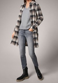 Camicia lunga in tartan nera, bianca e beige sopra un maglione grigio a scollo a V, abbinata a jeans skinny grigi e stivaletti neri con suola spessa.