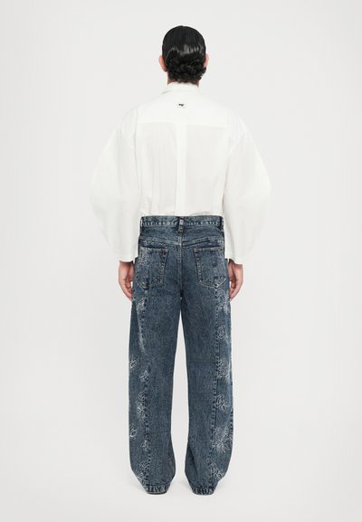 Camicetta bianca con maniche voluminose, abbinata a pantaloni di denim blu con ricami floreali e vestibilità rilassata. Vista da dietro.