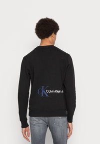 Černá mikina s kulatým výstřihem a vroubkovanými manžetami. Na zadní straně se nachází výrazné bílé a modré logo Calvin Klein Jeans.
