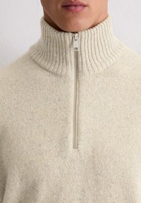 Beżowy sweter na zamek błyskawiczny z wysokim żeberkowanym kołnierzem, wykonany z fakturowanej dzianiny z subtelnymi niebieskimi i żółtymi drobinkami rozmieszczonymi w całej strukturze.