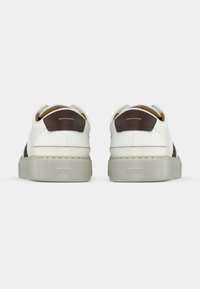 Sneakers bianche basse con dettagli marroni, punta rotonda e texture liscia. Presentano una suola spessa grigio chiaro e un dettaglio con logo sul tallone.