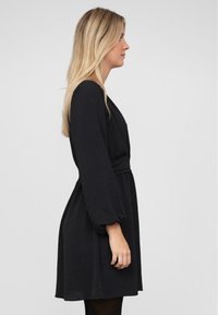 Vestido negro de manga larga con cintura entallada, que presenta un escote en V y mangas abullonadas. Fabricado en un tejido suave y texturizado.