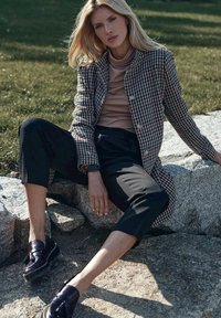 Płaszcz w wzór houndstooth w czerni i bieli, noszony na beżowym golfie, w połączeniu z czarnymi spodniami 3/4 i ciemnymi loafersami, siedzący na kamieniu.
