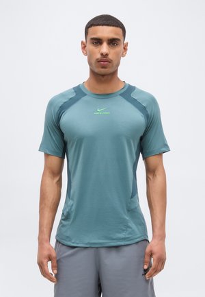 Νέος άνδρας στέκεται, φοράει τιρκουάζ αθλητική μπλούζα Nike Pro και γκρι σορτς, κοιτάζοντας εμπρός σε απλό φωτεινό φόντο.