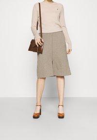 Haut en tricot beige à manches longues avec un petit emblème, associé à une jupe midi à carreaux marron. Sac bandoulière marron et talons compensés beige.