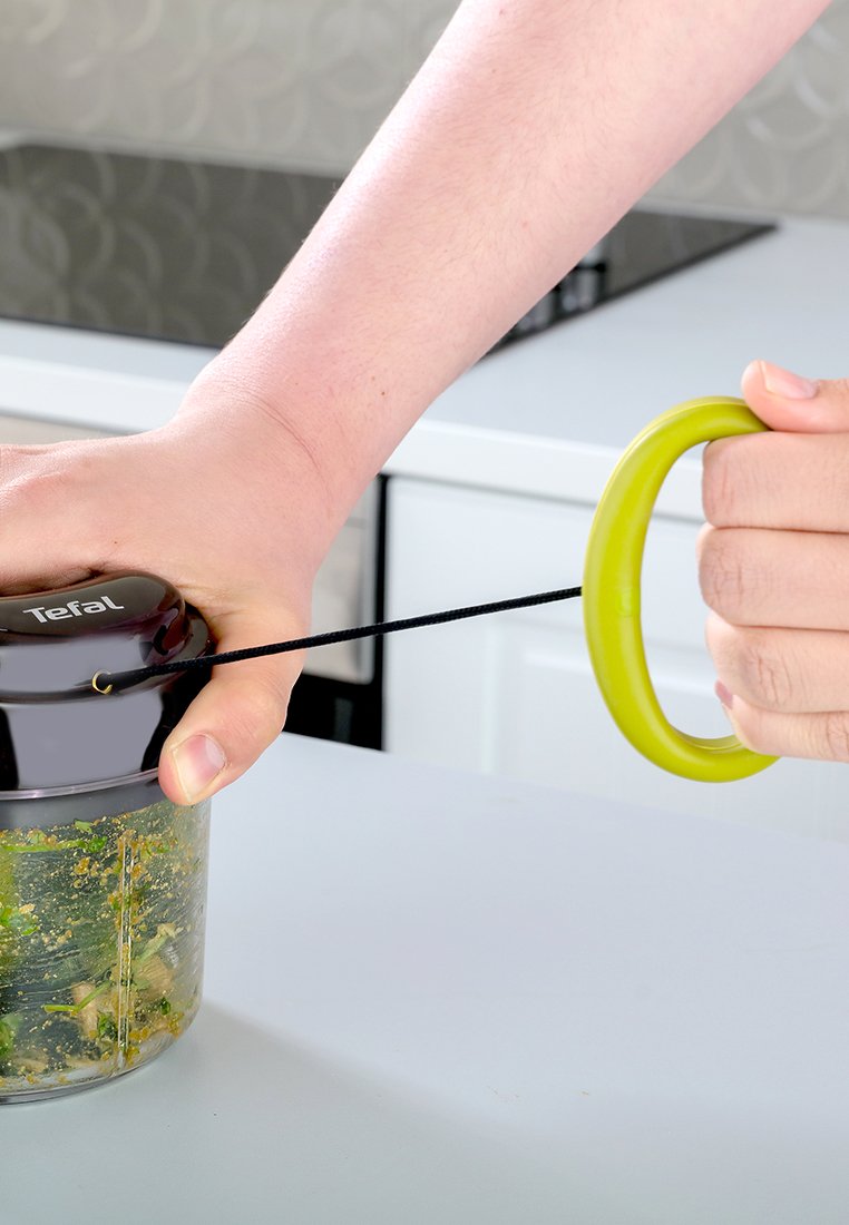 Hâchoir en plastique noir et transparent avec un manche vert. Une main tire sur une corde noire pour le faire fonctionner. Les contenus sont visibles à l'intérieur du récipient.