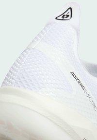 Valkoiset urheilukengät, joissa on teksturoitu verkkopinta, sileät korostukset ja kaareva pohja. Mukana kolmion muotoinen logo salaman kuvalla.