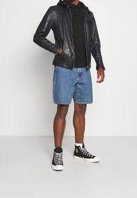 Mörk läderjacka med dragkedja, svart pikétröja, denimshorts och svarta högstövlar med vita sulor.