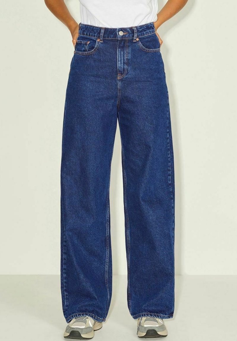 Breda blå denimjeans med hög midja, klassisk femfickdesign och subtila sömdetaljer. Stylist med grå sneakers.