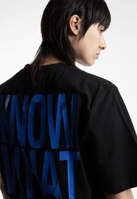 En person med kort sort hår vises fra siden, iført en sort T-shirt, der har store blå bogstaver, som siger "KNOW WHAT."
