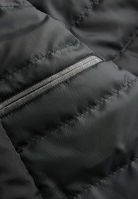 Sección de chaqueta de plumas negra con textura acolchada, líneas de costura horizontales y un bolsillo con cremallera oculta con una cremallera gris en el lateral.