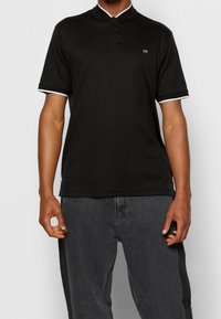 Polo noir à col montant, avec accents blancs sur les manches et un logo discret sur la poitrine. Confectionné dans un tissu lisse.