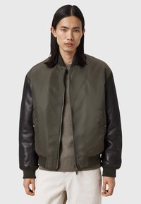 Homme aux longs cheveux noirs portant une veste bomber verte zippée avec des manches en cuir noir et un pantalon clair sur fond uni.