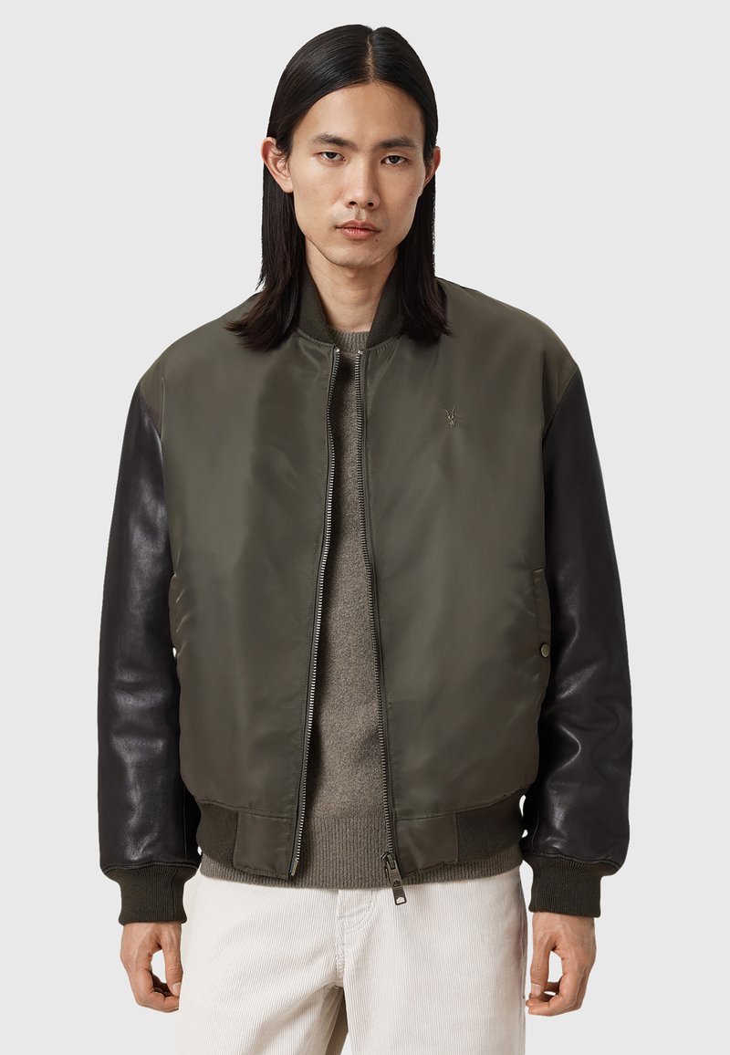 Homme aux longs cheveux noirs portant une veste bomber verte zippée avec des manches en cuir noir et un pantalon clair sur fond uni.