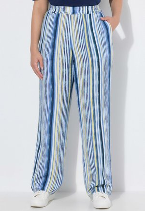 Person iført højtaljede, løse bukser med lodrette blå, hvide, gule og sorte striber, kombineret med hvide sneakers og en mørkeblå top.