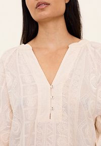 Light pink blouse featuring a V-neck, textured fabric, and decorative stitching with three buttons at the neckline.