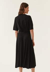 Schwarzes Kleid mit kurzen Ärmeln, lockerer Passform und fließendem Rock. Hergestellt aus weichem Stoff mit glatter Textur, verfügt über ein verdrehtes Rückendesign.