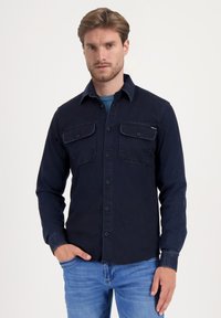 Camisa de manga larga azul marino de algodón con cuello abotonado, dos bolsillos en el pecho y un dobladillo ligeramente desgastado.