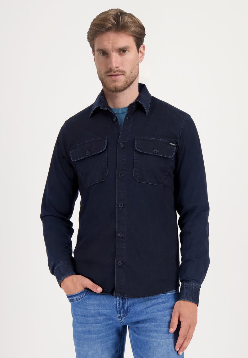 Camisa de manga larga azul marino de algodón con cuello abotonado, dos bolsillos en el pecho y un dobladillo ligeramente desgastado.