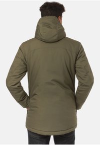 Hombre con chaqueta de invierno capucha color verde olivo, de pie con las manos en los bolsillos, mirando hacia el fondo blanco.