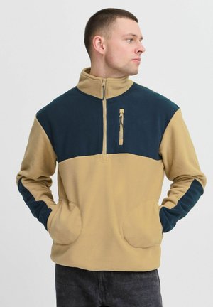 Fleece pullover with a zip collar, featuring a tan body and dark blue shoulders and sleeves. Includes zip pocket and side pockets.