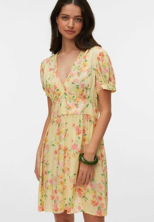 Mujer con vestido amarillo de flores, mangas cortas abullonadas y escote en V pronunciado, complementado con una pulsera verde en la muñeca.