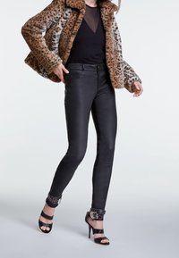 Persoon in een leopardprint bontjas, zwart nettop, hoge zwarte leren broek en zwart gestepte enkelbandhakken, staande in zijaanzicht.