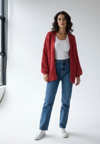 Czerwony sweter na guziki o fakturze, noszony na białym topie na ramiączkach, zestawiony z niebieskimi jeansami o prostych nogawkach i białymi trampkami. Miękka tkanina z otwartym przodem.