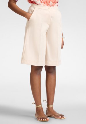 Frau trägt beige knielange Shorts, orange gemustertes Oberteil und goldene flache Sandalen mit Riemchen und Knöchelriemen.