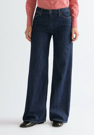 Osoba ubrana w ciemnoniebieskie szerokie dżinsy, czerwoną i białą koszulę w kratę oraz czarne buty, stojąca na tle jasnego, jednolitego tła.