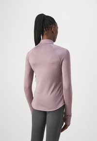 Femme avec des cheveux tressés portant un top mauve ajusté à manches longues et un legging gris foncé, debout de dos par rapport à la caméra.