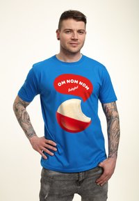 Blå kortärmad T-shirt med en stor röd pratbubbla med texten "OM NOM NOM Babybel" och en biten Babybel-ostgrafik.