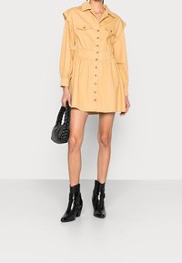 Lichtbeige overhemdjurkje met twee borstzakken, lange mouwen en een gegatherde taille; gecombineerd met zwarte enkellaarsjes en een gevlochten tas.