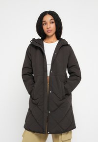Ung kvinde iført en lang sort quiltet frakke med hætte, en hvid cropped top og beige cargo-bukser, stående med hænderne i lommerne.