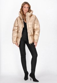 Złota kurtka puchowa z wysokim kołnierzem, pikowany design i zamek błyskawiczny; zestawiona z czarnymi dopasowanymi spodniami i czarnymi botkami na obcasie.