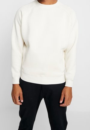 Homme portant un sweat-shirt blanc uni à col ras du cou et un pantalon noir, debout avec les bras détendus le long du corps devant un fond blanc.