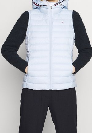 Personne portant une doudoune sans manches bleu clair avec capuche, un pull noir à manches longues en dessous et un pantalon noir, mains dans les poches de la veste.