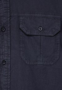 Tmavě modrá košile s knoflíky vyrobená z lehkého materiálu, která má náprsní kapsu s šikmým klopou a černými plastovými knoflíky.