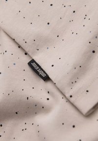 Beige katoenen stof met een gevlekte patroon in zwart en blauw. Heeft een zwarte geweven label met witte tekst, gepositioneerd bij een naad.