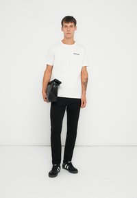 Hvid t-shirt med "REPLAY" tekst, sorte bukser og sorte sneakers med hvide detaljer. Model der holder en sort taske, neutral baggrund.