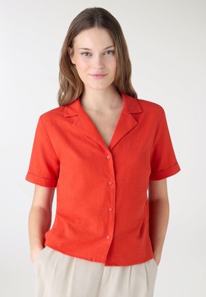 Giovane donna con capelli castano chiaro che indossa una camicia arancione a maniche corte con bottoni e pantaloni beige, in piedi davanti a uno sfondo semplice.