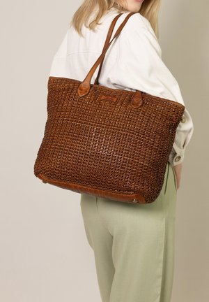 Geflochtene braune Ledertasche mit zwei Trägern, die einen strukturierten Körper und einen glatten Boden aufweist, präsentiert an einer Person in lässiger Kleidung.