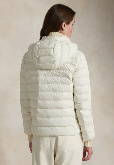 Vrouw draagt een crèmekleurige doorgestikte pufferjas met capuchon, gecombineerd met bijpassende lichte broek, gezien van achteren.
