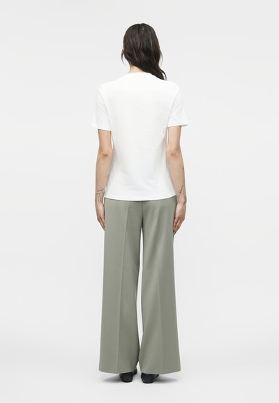 Γυναίκα με σκούρα μαλλιά που φορεί λευκή μπλούζα με κοντά μανίκια και άνετο φαρδύ παντελόνι σε απόχρωση φασκόμηλου, όρθια με την πλάτη γυρισμένη σε λευκό φόντο.