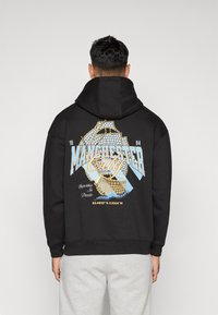 Musternööriga must kapuutsiga sviiter, millel on graafiline disain koos "Manchester City" tekstiga, koordinaatidega ja sinise ja kollase värvi aktsentidega.