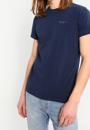 Osoba ubrana w granatowy t-shirt z krótkim rękawem z jasnym logo na piersi i jasnoniebieskie dżinsy, stoi z ramionami swobodnie opuszczonymi wzdłuż ciała.