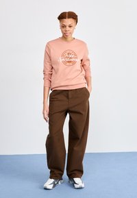 Lys rosa hettegenser med "Nike Sportswear" grafikk, paret med løse brune bukser og grå-hvite sneakers. Glatte stoffteksturer.